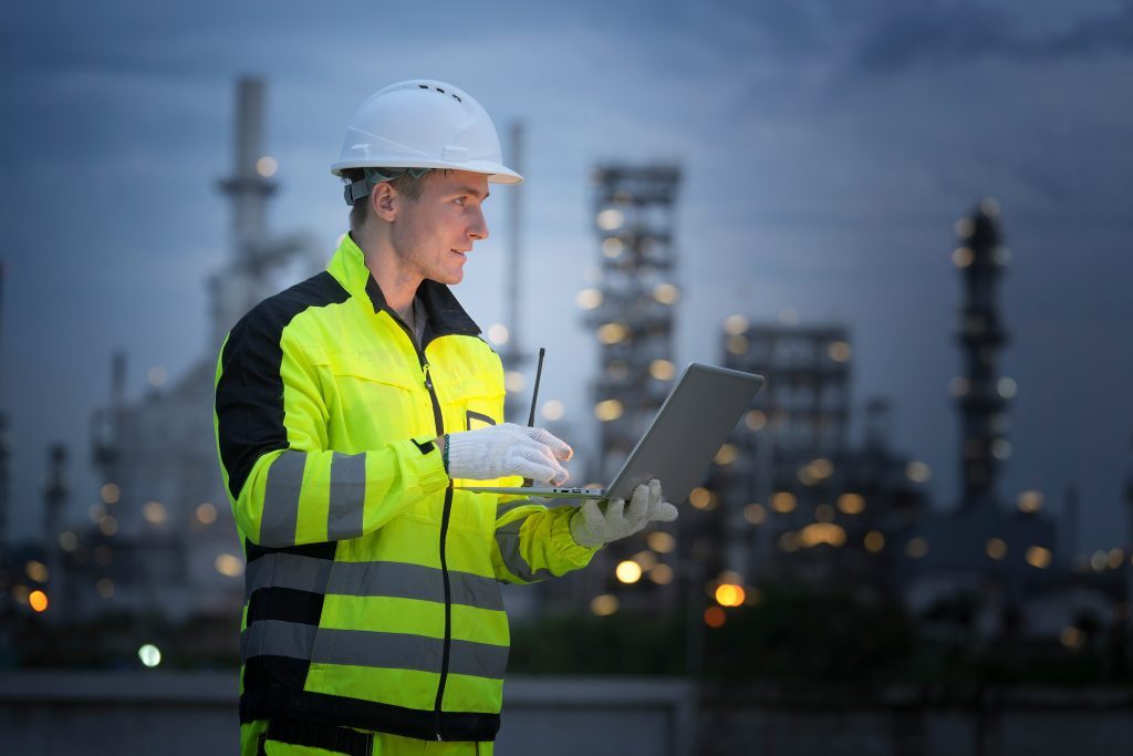 A worker in a bright yellow safety suit stands in an industrial area, focused on a laptop. The background features tall refinery structures illuminated against the night sky.